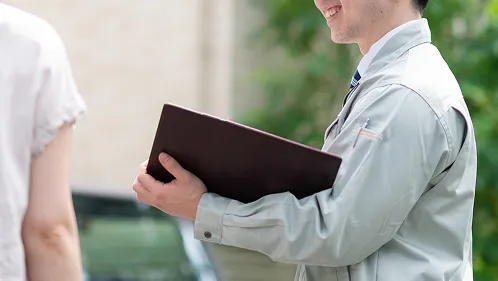グレーの作業着にストライプ柄ネクタイの男性スタッフが、茶色のバインダーを手に笑顔で対話している様子