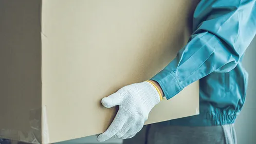 水色の作業着に白い作業手袋をした人物が大きなダンボール箱を抱えている手元のクローズアップ