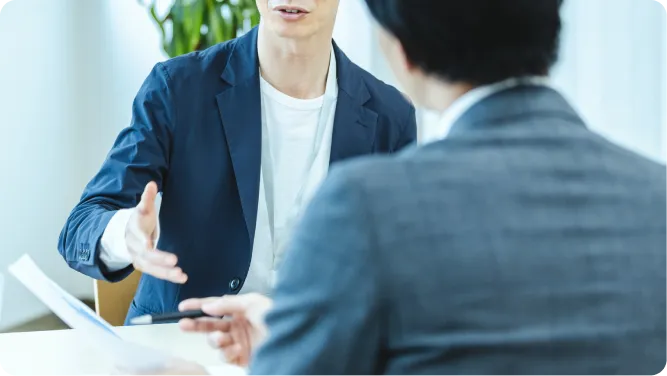 紺色のジャケットを着た男性が書類を手に持ちグレースーツの相手と向かい合って打ち合わせをしている様子