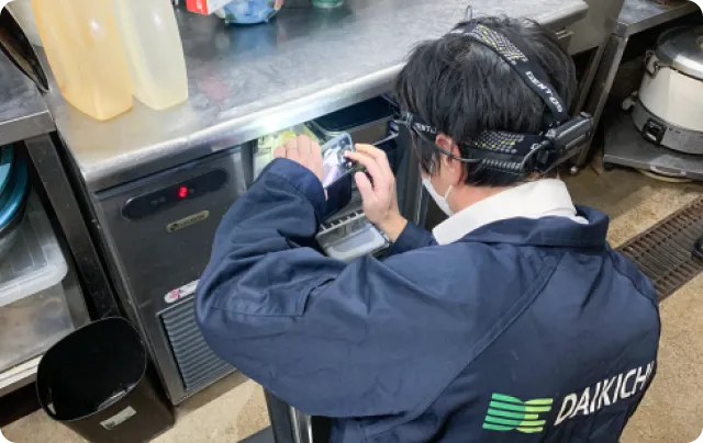 厨房でヘッドライトを装着し、業務用冷蔵庫の内部を点検する作業スタッフの後ろ姿